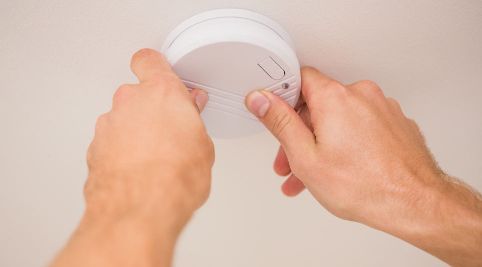 Two human hands operating a white smoke alarm on a ceiling