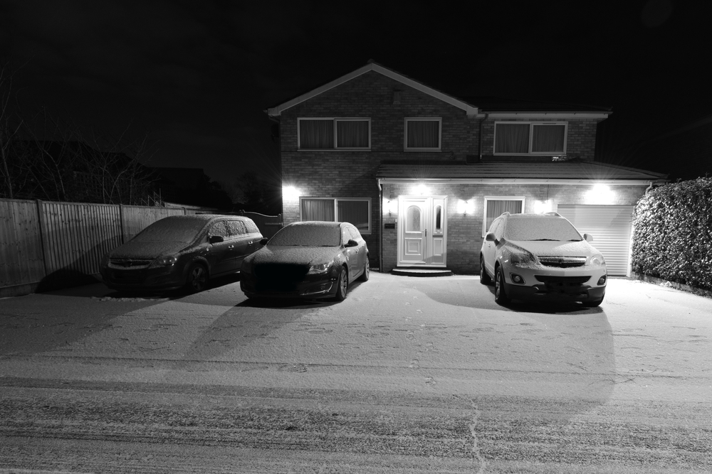 CCTV at night time capturing snowy road with a lit up house and three cars in the driveaway